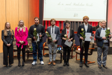 Siedem osób pozuje do zdjęcia. Wszyscy są elegancko ubrani. Pięć osób trzyma w dłoniach po jednym kwiecie oraz dyplom, dwie mają dodatkowo kryształowe statuetki.