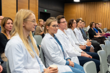 A group of people are seated in an auditorium during a formal event. In the front row are people wearing white coats, likely medical professionals or scientists, followed by attendees in business attire. The room has wood paneling on the walls and rows of gray chairs.