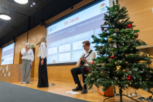 A scene during the celebrations of the 55th anniversary of the Clinic of Pediatric Oncology, Hematology, and Clinical Transplantology. On the left, two people are standing, one singing into a microphone, and on the right, a person is sitting and playing the guitar. In the background, there is a screen with an inscription about the anniversary and a decorated Christmas tree.