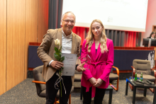 Dwie elegancko ubrane osoby pozują do zdjęcia. Mężczyzna po lewek trzyma w ręce kwiat i dyplom. Obie osoby uśmiechają się do zdjęcia. 