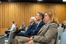 Kilka osób siedzi w sali z drewnianymi panelami na ścianach, uważnie obserwując wydarzenie. W pierwszym planie widoczne są trzy osoby, a w tle pozostali uczestnicy.