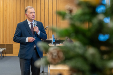 A man in a suit speaks into a microphone in a conference room with wooden paneling and chairs in the background. A blurred Christmas tree is visible in the foreground, suggesting that the event is taking place during the holiday season.