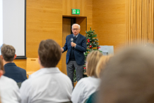 A man speaks into a microphone in front of an audience in a room with wooden walls. A decorated Christmas tree is visible in the background, suggesting that the event is taking place during the holiday season.