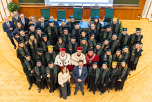 A large group of people stand in a semicircle in a lecture hall with wooden floors and light-colored walls. Most of them are dressed in black gowns with green trim and black caps, while a few have yellow or blue caps. In the center are people in rector's robes with red caps and decorative chains.