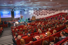 Sala audytoryjna jest wypełniona uczestnikami siedzącymi na czerwonych fotelach. Na scenie stoją dwie osoby przy mównicy, obok której znajdują się trzy choinki ozdobione białymi bombkami i lampkami. Na suficie widoczne są projekcje śnieżynek, a na dwóch dużych ekranach wyświetlają się slajdy z tekstem w kolorze białym na niebieskim tle. 