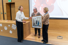 Three people are standing in a room with wooden walls, next to a large screen showing a presentation. One person is handing a framed photo to another, while the third person is standing nearby.