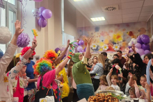 A group of people, adults and children, dressed in colorful clothes, are celebrating in a large, bright room. There are purple balloons around them, and drawings on the walls.