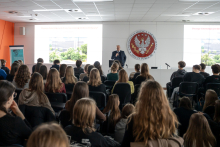 Prof. Czupryniak w czasie wykładu stoi na środku sali. Za nim widać czerwone godło WUM oraz ekran multimedialny z wyświetlonymi zdjęciami. Przed nim w rzędach siedzą młodzi ludzie.