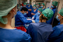 A group of people during a medical procedure. They are all wearing blue gowns, caps on their heads, and masks on their faces. In the center, two people are sitting at a treatment table.