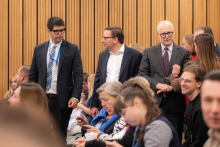 Na zdjęciu znajduje się grupa osób w sali konferencyjnej lub audytorium. W tle widoczne są pionowe drewniane panele na ścianie. W centralnej części kadru stoją trzy osoby ubrane w formalne stroje – garnitury z krawatami i koszulami.