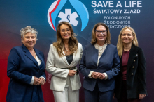 Four women stand in front of a wall with the inscription “SAVE A LIFE – World Congress of Polish Medical Communities.” They are all dressed elegantly. The background of the wall is navy blue and red with a large logo at the top.