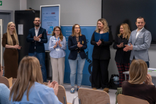 Siedem osób stoi w rzędzie w sali konferencyjnej. Osoby trzymają dłonie złożone lub klaszczą. W tle widać baner uczelni i ekran z informacją o wydarzeniu. Przed nimi siedzi kilka osób na krzesłach, które również klaszczą.