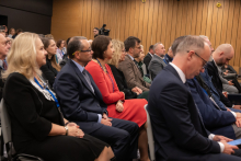 Na zdjęciu widać osoby siedzące w rzędach na sali konferencyjnej. W tle znajdują się drewniane panele na ścianie, a w głębi kadru widoczny jest operator kamery. Uczestnicy mają na sobie eleganckie stroje – garnitury, marynarki oraz sukienki,