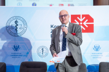 An elegantly dressed man, Prof. Mariusz Gujski, stands with a microphone in his hand and speaks. In the background, you can see a wall with the event logos.