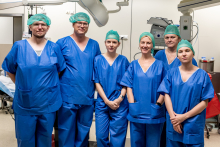 Six people dressed in blue medical uniforms and green caps pose for a photo in an operating room. Medical equipment can be seen in the background.