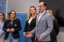 Pięć osób stoi obok siebie w sali. Osoba z prawej strony gestykuluje rękami, jakby tłumaczyła lub prezentowała. Pozostałe osoby stoją spokojnie, trzymają ręce przed sobą. W tle widoczny jest niebieski baner uczelni i fragment ekranu.