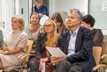 Grupa osób siedzi na krzesłach w jasnym pomieszczeniu przypominającym salę konferencyjną. W pierwszym rzędzie mężczyzna w garniturze trzyma dokumenty, obok niego kobieta w czarnej marynarce i czerwonej sukience. W tle widać kolejne osoby