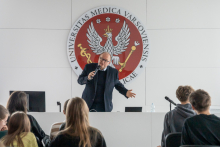 Prof. Czupryniak w czasie wykładu stoi na środku sali. Za nim widać czerwone godło WUM. Przed nim w rzędach siedzą młodzi ludzie.