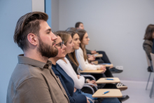 Siedem osób siedzi w jednym rzędzie w sali. Każda osoba patrzy w stronę przodu, gdzie prawdopodobnie odbywa się prezentacja. Na małych stolikach przed nimi leżą długopisy. 