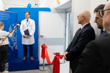 A person in a white doctor's coat stands on a raised platform in front of a blue wall with the words “Warsaw Medical University” written on it, holding a navy blue briefcase in their hands. They speak into a microphone on a stand. On the left is a person with a microphone and a smartphone.