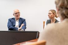 An elegantly dressed man during a meeting. He is explaining something, gesturing with his hand. Next to him, on the left, a young woman is sitting.