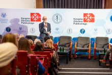 An elegantly dressed man, Dr. Tomasz Maciejewski, stands with a microphone in his hand in the middle of the auditorium. Behind him is a wall with the event logos, and in front of him, the congress participants are seated in rows.