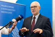 A person in a dark suit with a burgundy tie stands in front of a blue wall with the words “Warsaw Medical University.”  He gestures.