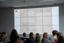 Ekran multimedialny ze zdjęciem starego dokumentu oraz młodzi ludzie siedzący na krzesłach i patrzący na ten dokument.