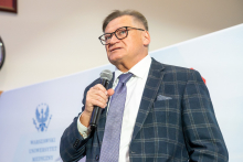 An elegantly dressed man, Prof. Bolesław Samoliński, stands with a microphone in his hand and speaks. In the background, you can see a wall with logos.