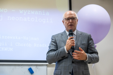 An elegantly dressed man, the rector of the Medical University of Warsaw, speaks into a microphone. In the background, there is a multimedia screen and a purple balloon.