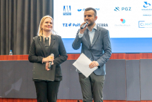 Dwie osoby, kobieta i mężczyzna, elegancko ubrani, stoją z mikrofonami w rękach. Mężczyzna mówi do mikrofonu. W tle widać ekran multimedialny.