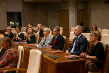 Grupa osób siedzi w eleganckiej sali konferencyjnej przy drewnianych stołach z kremowymi fotelami. Na stołach leżą broszury i notatniki, część osób je trzyma.