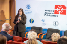 An elegantly dressed woman, Monika Kowalczyk, stands with a microphone in her hands and speaks. Behind her, you can see a wall with logos. In front of her, congress participants sit in rows.