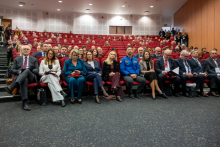 A large group of people are sitting in red chairs in an auditorium during a conference. 
