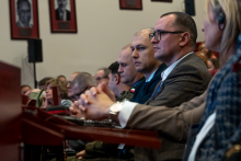 A group of elegantly dressed people are sitting in the auditorium on red chairs. They are looking straight ahead, focused.