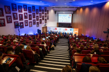A shot of the auditorium filled with congress participants. At the bottom of the auditorium, you can see the panelists sitting in chairs. Behind them is a wall with logos and a multimedia screen.