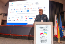 A man standing behind a lectern. In the background, there is a multimedia screen with many different colored logos. On the right, there are three flags: Ukraine, Poland, and the European Union.