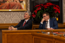 Dwóch elegancko ubranych mężczyzn siedzi przy drewnianym stole w sali obrad; na ścianie widoczny obraz w ramie, a w tle okno ozdobione dużą dekoracją z czerwonych poinsecji