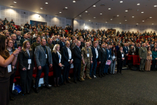 A very large group of people standing in a large auditorium. They are listening to the national anthem.