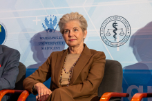 An elegantly dressed woman is sitting in an armchair. Behind her, you can see a wall with logos.