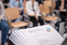 Stos papierów z wydrukowanym tekstem leży na pierwszym planie. Na górnej kartce widoczny jest tytuł dotyczący olimpiady wiedzy oraz lista pytań testowych. W tle kilka osób siedzi na krzesłach w sali, trzyma notatki lub długopisy. 