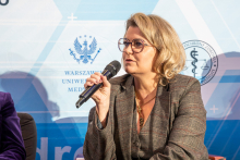 An elegantly dressed woman is sitting in an armchair. She is holding a microphone in her right hand and speaking into it. In the background, there is a wall with logos.