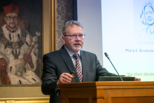 Mężczyzna w garniturze stoi za drewnianą mównicą z mikrofonem, trzymając pilot do prezentacji. W tle widoczny ekran ze slajdem zawierającym imię „Mary E. Brunkow” oraz fragment cytatu