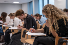 Kilka osób siedzi w sali i pisze na kartkach. Każda osoba trzyma długopis i wypełnia dokumenty na małych stolikach przymocowanych do krzeseł. Widać skupienie na pisaniu testu.