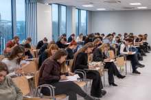 Około trzydzieści osób siedzi w dużej sali przy oknach. Każda osoba pisze na kartkach, trzyma długopis i korzysta z małych stolików przymocowanych do krzeseł. Wszyscy są skupieni na pisaniu testu.