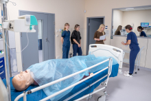 Cztery osoby znajdują się w sali medycznej. Jedna osoba stoi przy łóżku i rozmawia z osobą widoczną w lustrze, druga trzyma kartki, a dwie stoją obok drzwi. Na łóżku leży fantom przykryty niebieskim materiałem. W tle widać sprzęt medyczny i monitor z parametrami. 