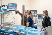 Trzy osoby stoją przy łóżku w sali medycznej. Na łóżku leży fantom przykryty niebieskim materiałem. Jedna osoba wskazuje ręką na monitor z kolorowym tekstem, który jest częścią symulacji. Pozostałe osoby patrzą na ekran i słuchają. W tle widać sprzęt medyczny i aparaturę.