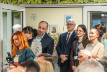 Grupa osób znajduje się w jasnym pomieszczeniu podczas oficjalnego wydarzenia lub prezentacji. Na pierwszym planie siedzą uczestnicy trzymający dokumenty, a w tle kilka osób stoi przy drzwiach i fotografuje lub nagrywa przebieg spotkania smartfonami i kamerami.