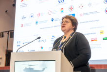 An elegantly dressed woman stands behind a lectern. She speaks into a microphone. In the background, there is a multimedia screen displaying many different colored logos.