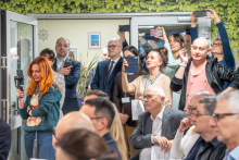 Grupa osób znajduje się w jasnym pomieszczeniu podczas oficjalnego wydarzenia lub prezentacji. Na pierwszym planie siedzą uczestnicy trzymający dokumenty, a w tle kilka osób stoi przy drzwiach i fotografuje lub nagrywa przebieg spotkania smartfonami i kamerami.